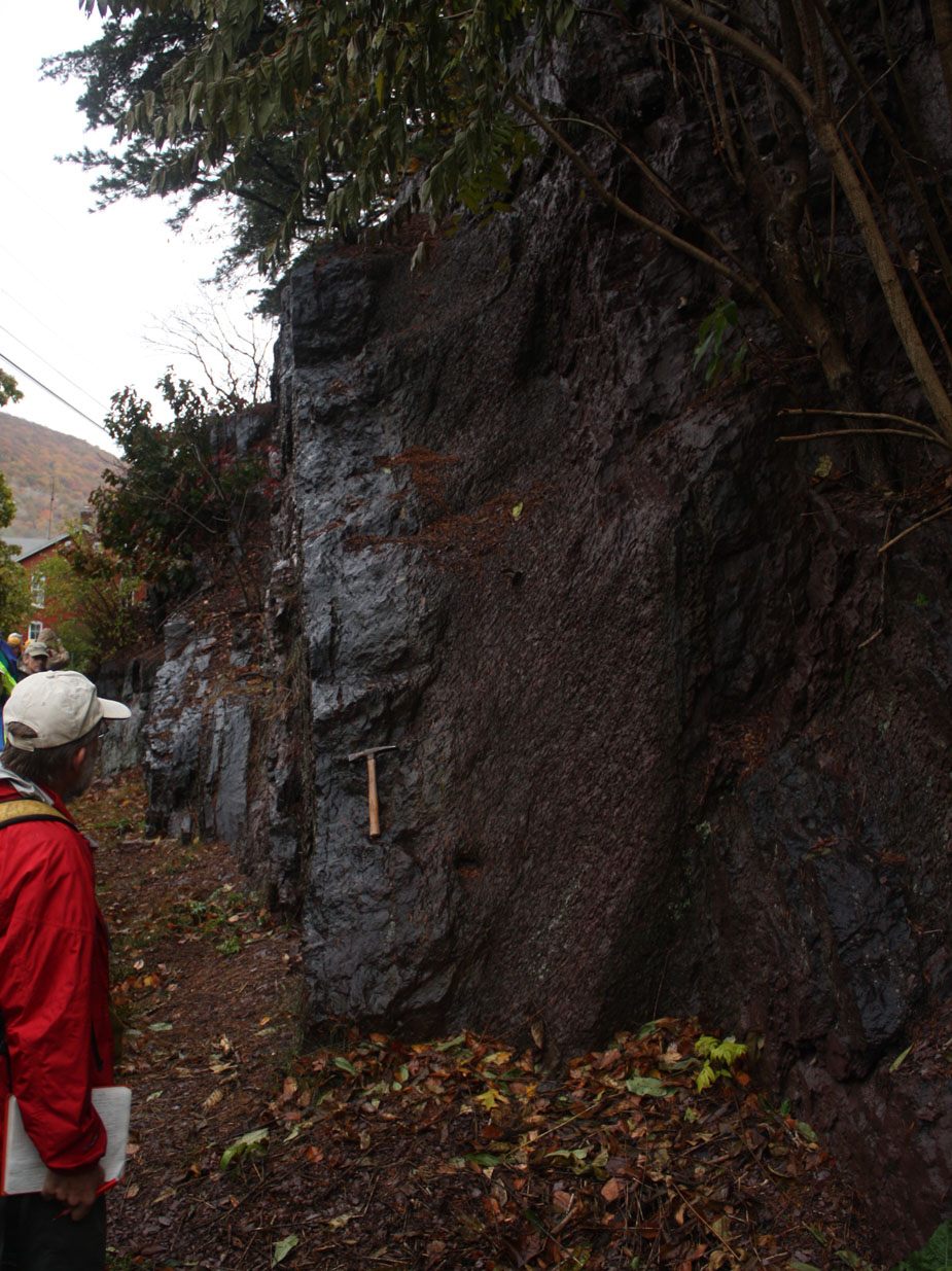 A dark, blocky rock face with a geologist's hammer wedged into a crack.