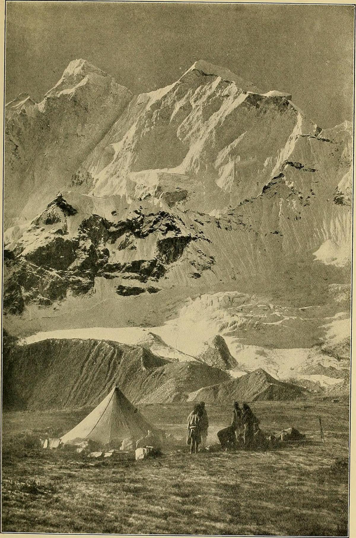 A sepia photograph of a canvas tent and three figures camped beneath a snow-covered mountain face.