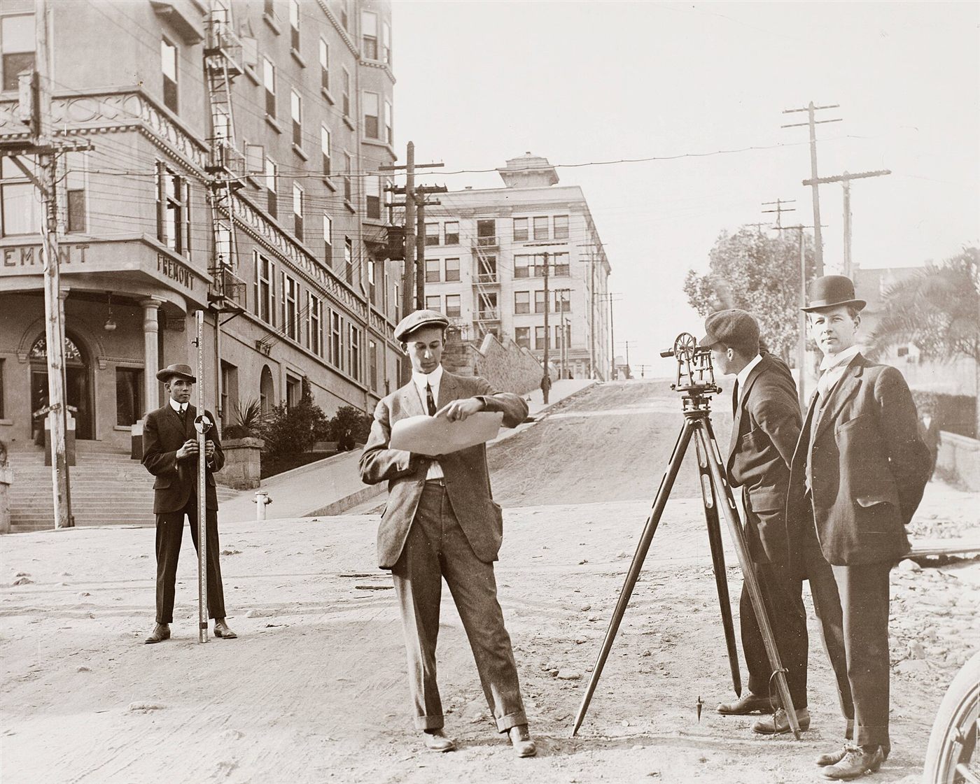 Three surveyors in suits with a transit on a tripod and a levelling staff on a city street.